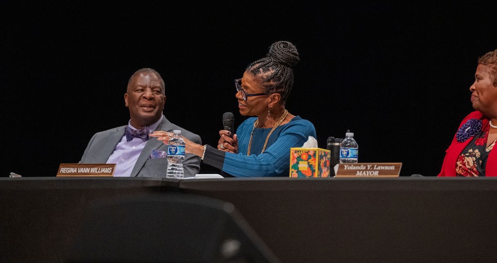 Panel of three at a formal event: a man in a gray suit with a bow tie on the left, a woman in blue center speaking into a microphone, and a woman in red on the right; nameplates and water bottles are on the table.