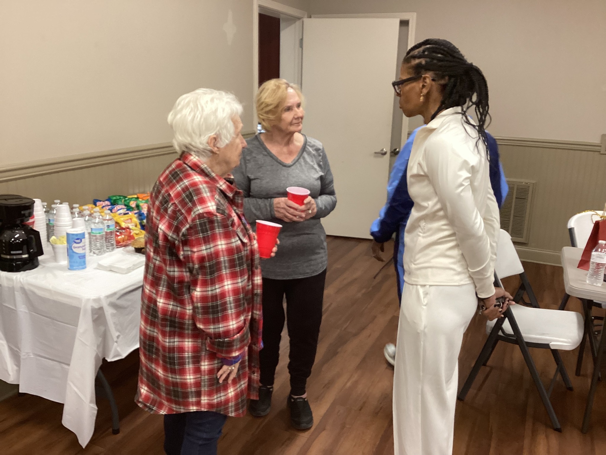 Two women talk near a snack-and-drink table; one wears a red plaid shirt and the other a gray top, with a nurse in white nearby.