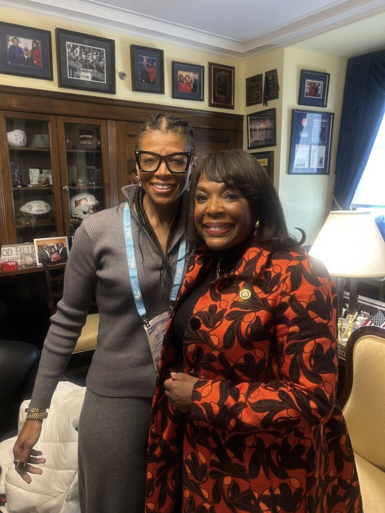 Two women smiling for a photo in an office, with framed pictures and awards on the walls behind them.